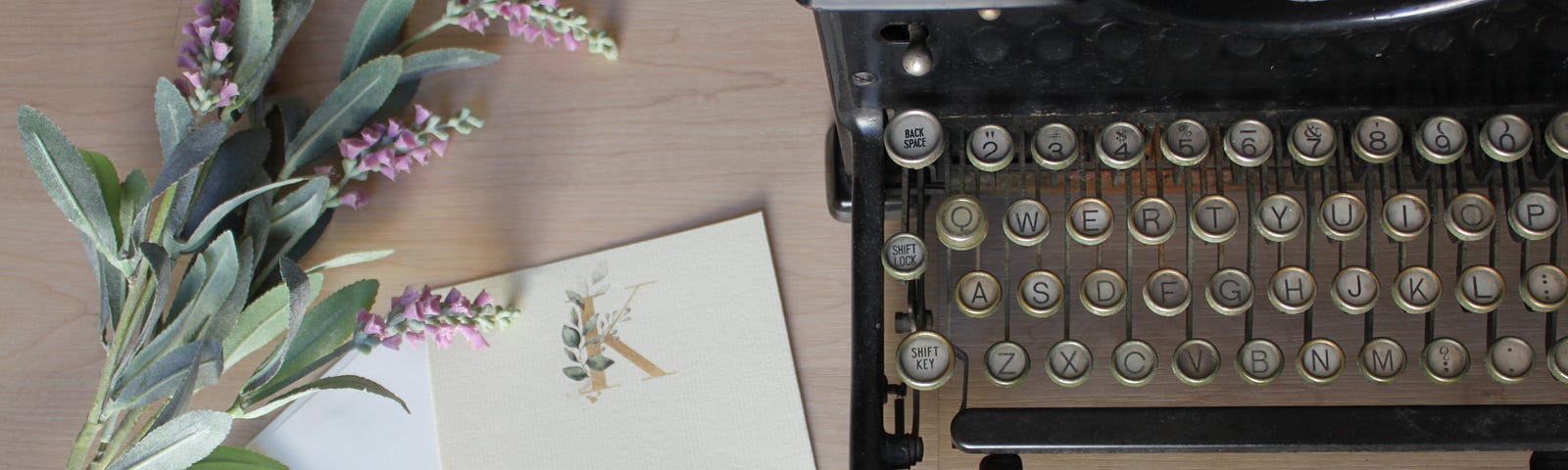 An antique typewriter sits on a wooden desk with a pad of paper and a sprig of purple flowers.