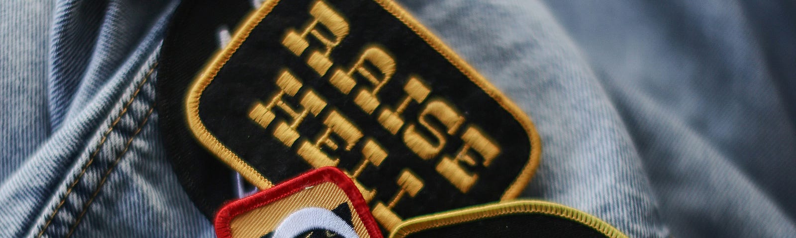 This image shows a collection of quirky and colorful patches on a denim jacket. The patches feature various fun and humorous messages such as “Raise Hell”, “I’m Not Tired”, and “Support Your Local Emotional Human”. The patches vary in shape and color, adding a personalized touch to the jacket.