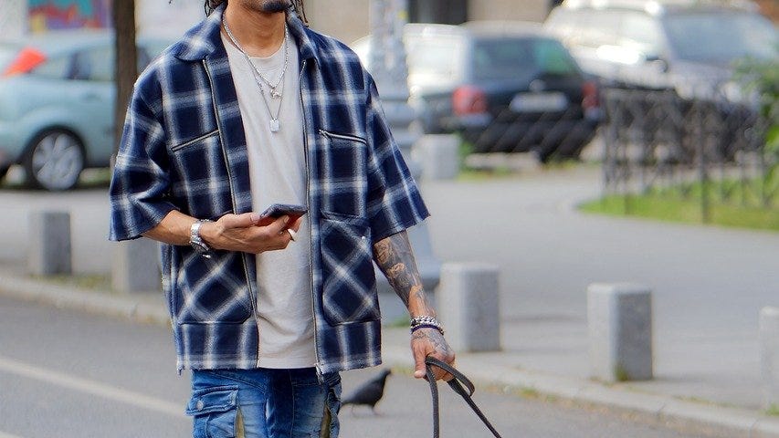 Brown-skinned man in his 30s, dreadlocks and goatie with black glasses and a white beanie, wearing white headphones, a white shirt, two necklaces, a blue and white flannel, and blue jeans with white sneakers. He’s holding a cellphone in his right hand while walking a blonde dog with a red bandana on a city street.