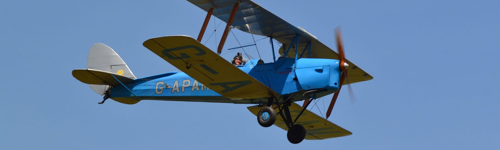 Old-fashioned bi-plane with propeller flying against blue sky. Pilot’s head visible.