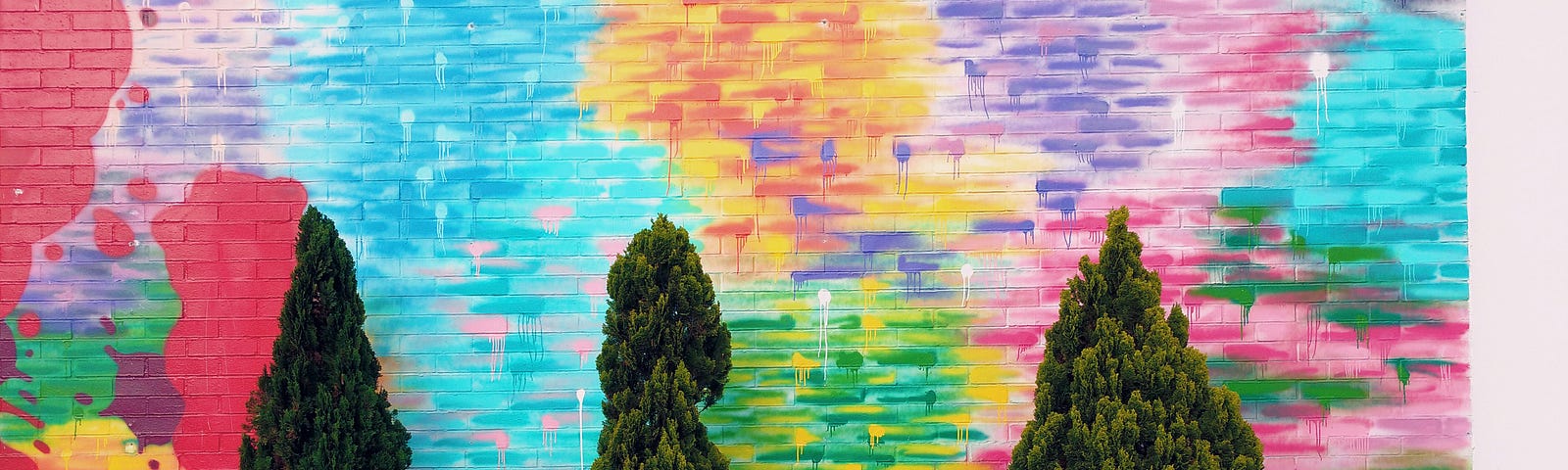 The image shows a vibrant, colorful mural on a wall, with the artwork giving the impression of an abstract sunset with blue sky above and various colors blending into each other. In front of this mural, there are real, three-dimensional shrubs and plants, which include several conical trees and a row of bushy plants with pink flowers, adding a layer of life in contrast to the painted wall. This juxtaposition of living plants against the wall creates a playful interplay between art and nature.