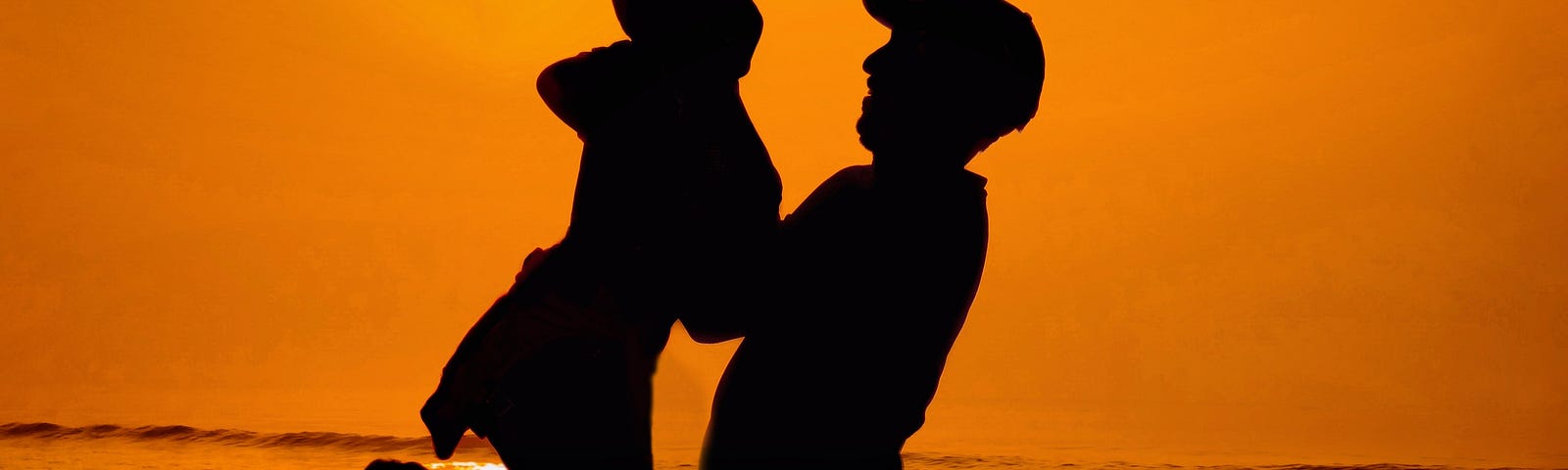 Father wearing a baseball cap holding a child in the air on the beach during a sunset