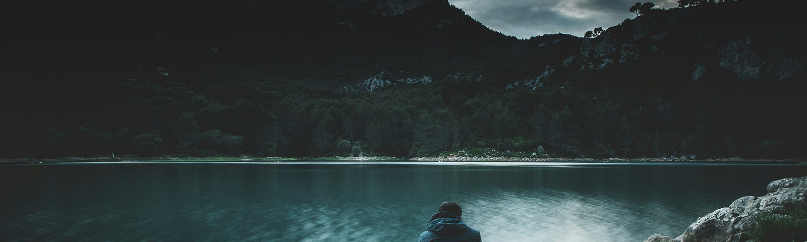 A man sitting buy a lake with his back turned and staring at the lake and the hills in the background.