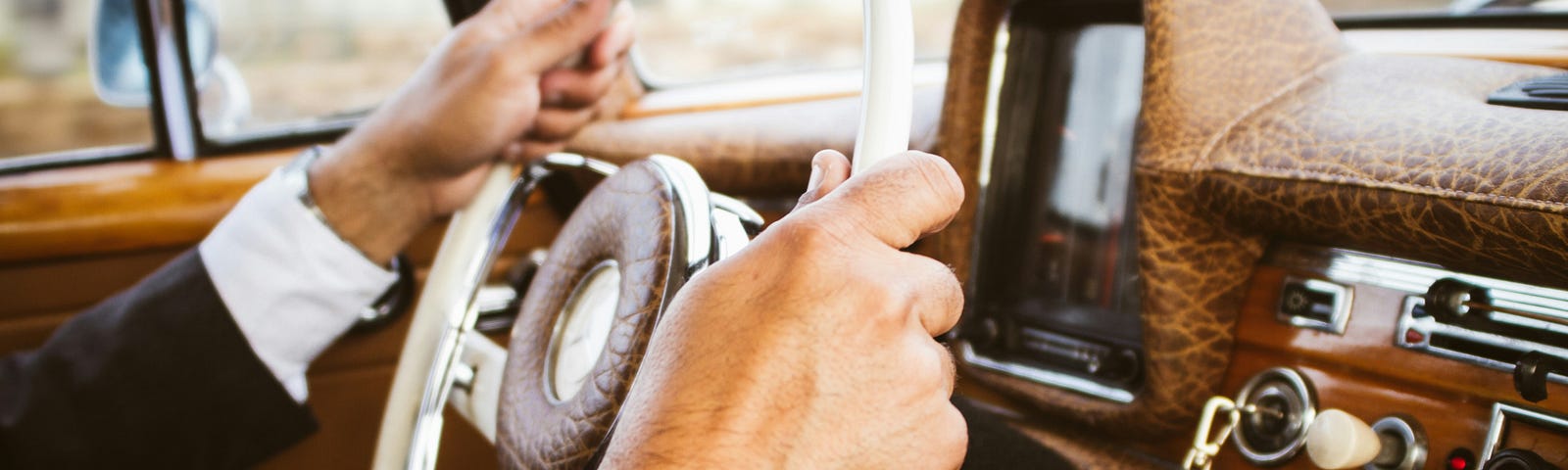 Man in a suit and dress shirt, grips the steering wheel of an old classic car, key in the ignition.