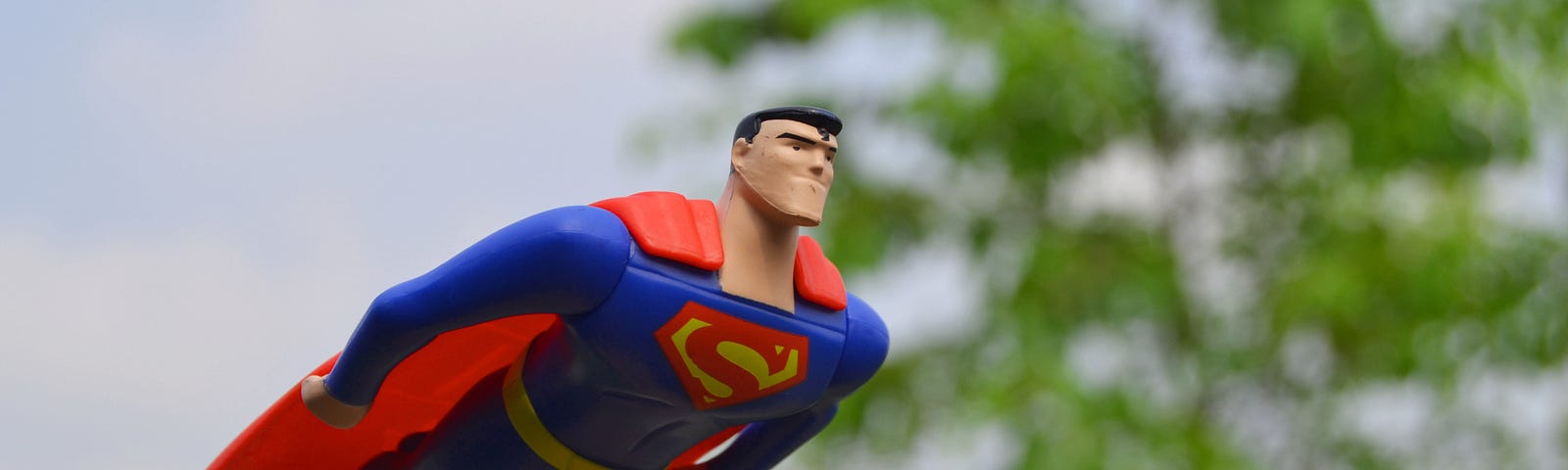 An action figure of Superman flies through the air in front of a green tree