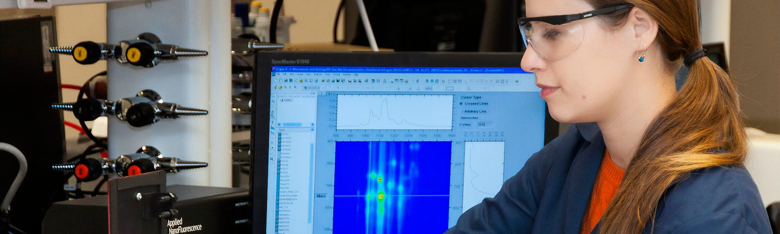 A woman in safety goggles holds a vial before a computer monitor.