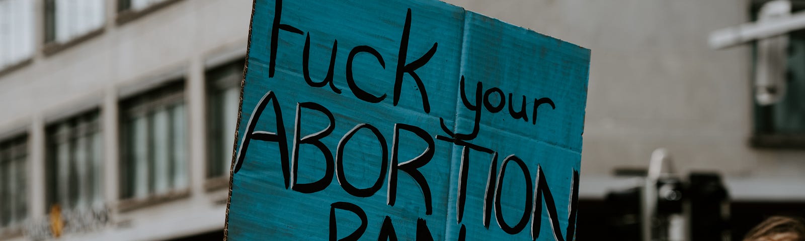 A protest sign, blue with black lettering reading, “Fuck your Abortion ban”