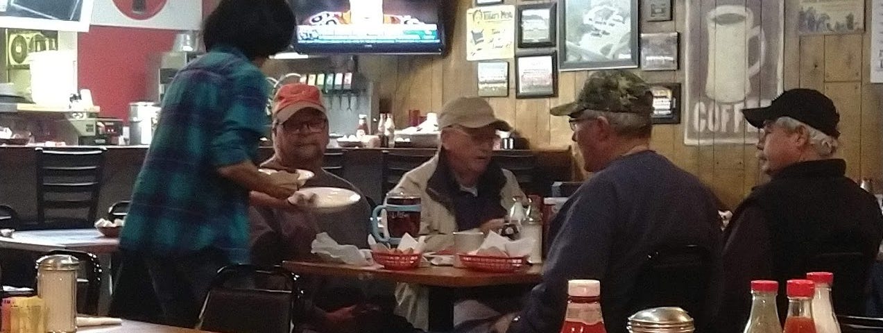 A waitress at a diner serves a group of older men gathered around a table