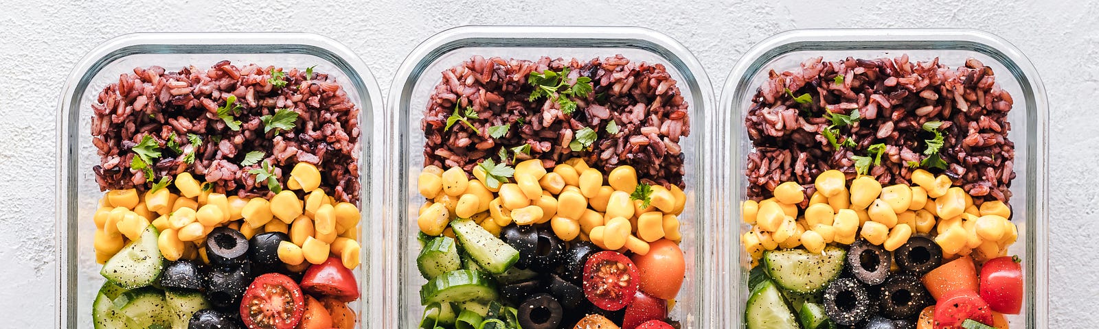 looking down on a white counter with 3 glass containers of perfectly prepared food, all 3 look identical with a combination of greens, rice, corn, tomatoes, olives, cucumber and mushrooms.