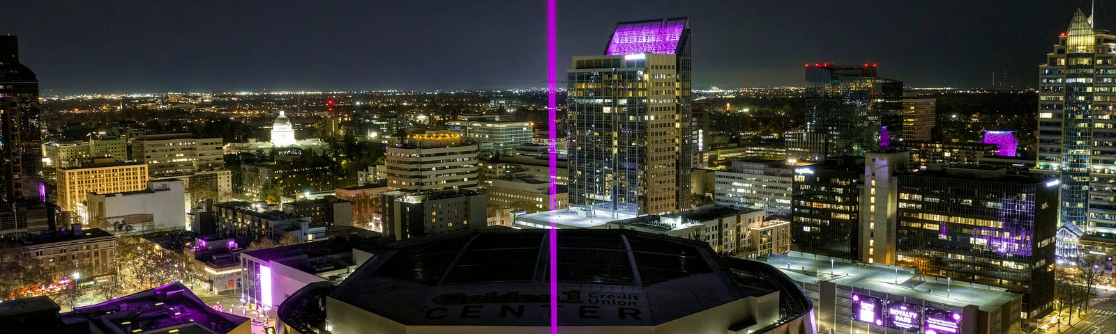 A bright purple beam shooting into the sky from the Sacramento Kings arena in Sacramento, California, surrounded by buildings