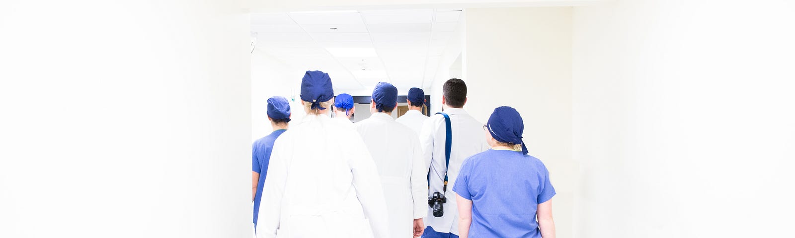 A group of doctors, nurses and anaesthetists wearing blue scrubs and white gowns, walk along an endless white corridor