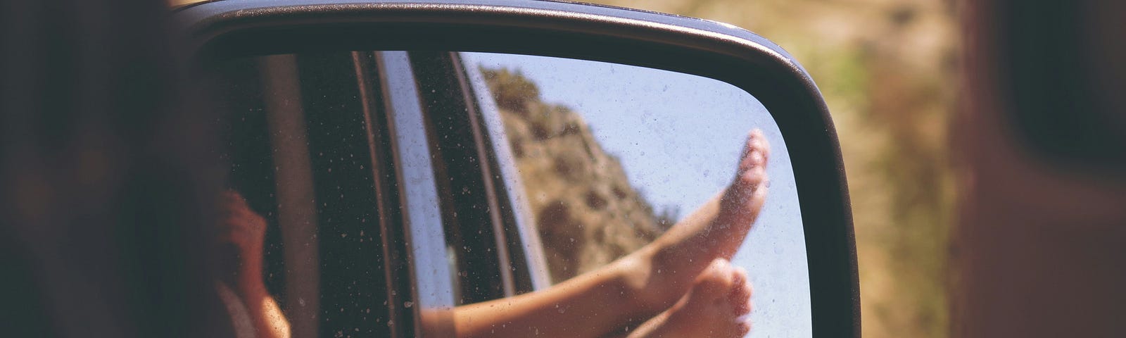 A reflection of bare feet sticking out of a moving car window, captured in the side mirror during a sunny drive through a rural area.