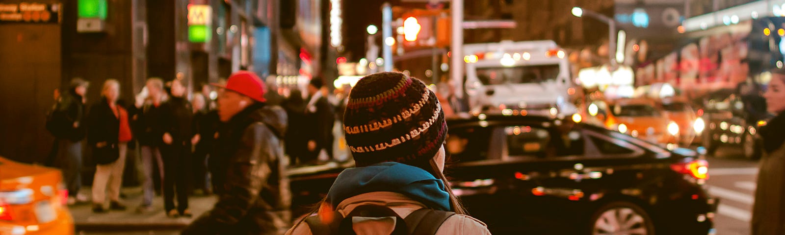 A busy city street at night. Cars are driving by. A lone girl waits to cross the street.