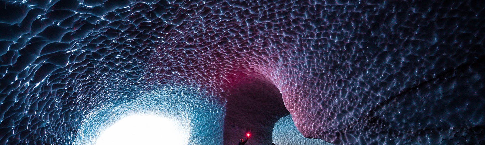 Inside a mysterious cavern looking out to the brightly lit entrance with the interior ceilings dimpled in deep waves throughout. A person is inside holding a reddish light over their head making the ceiling a pinkish hue just above them.