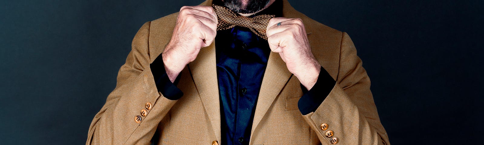 A bearded man in a brown suit, grey hatand black shirt balances a green apple on his head as he adjusts his fancy bowtie.