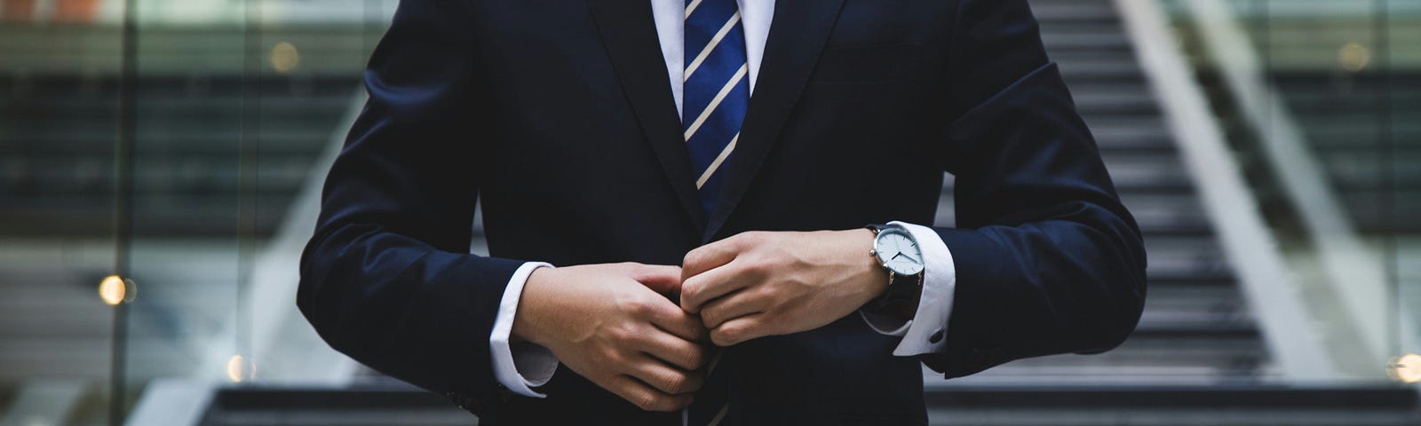 Man in a blue business suit with a white shirt and blue-and-white striped tie buttons his jacket.