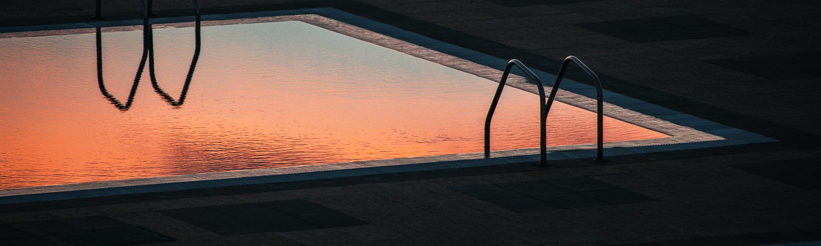 A sunset reflecting off the surface of a pool. The reflection of the ladder adding a plot twist. One ladder allows you to climb out of the water, the other seems to guide you in, deep below the surface.