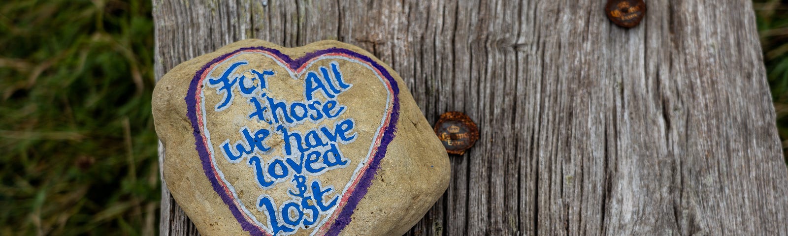 A rock sits on a rough piece of grey wood. On the rock is a painted heart and the words “For all those we have loved & lost.”