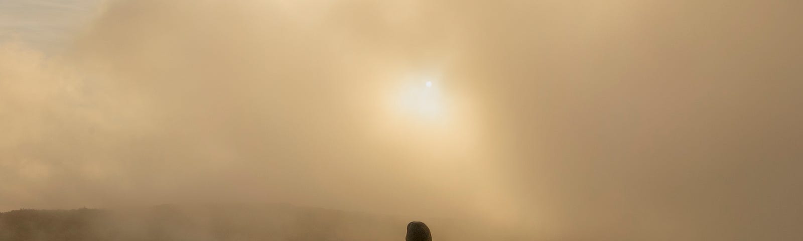 A person sits on a rocky outcropping, their back to the viewer. The air is foggy, the sun filtering through.