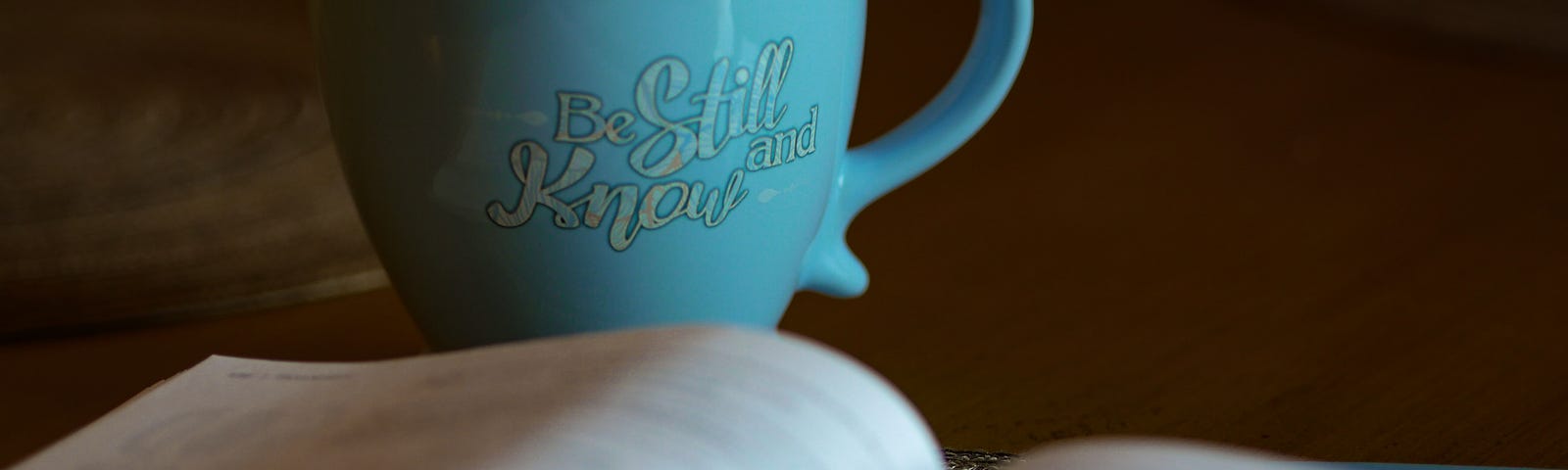 Teal coffee cup that says “Be Still and Know” next to an open bible on a table.