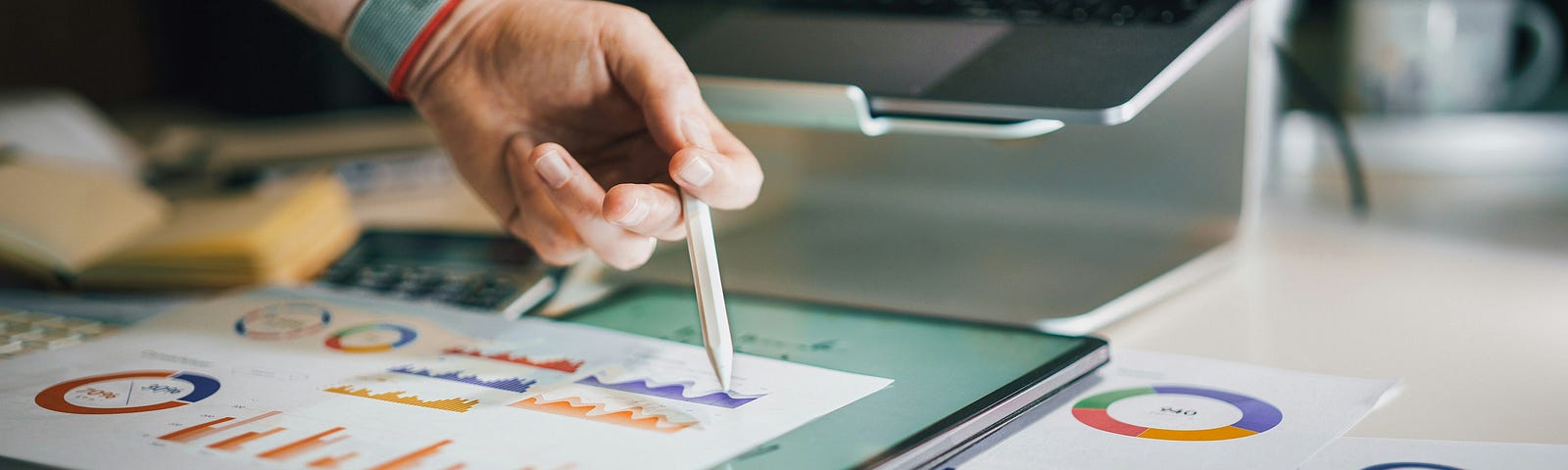 A person analyzing data charts and graphs on a tablet and paper, surrounded by a laptop and calculator, highlighting the power of augmented analytics in business.