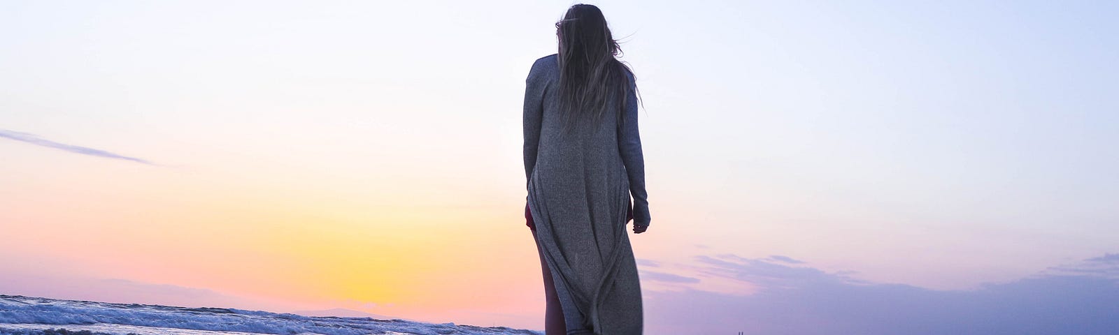 woman back toward us wearing long coat walking along beach sunrise