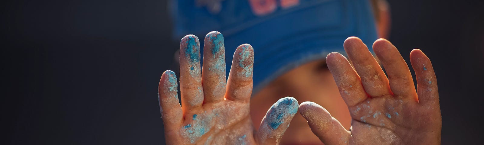 The messy, painted small hands of a young boy, pre-school aged, wearing a ball cap.