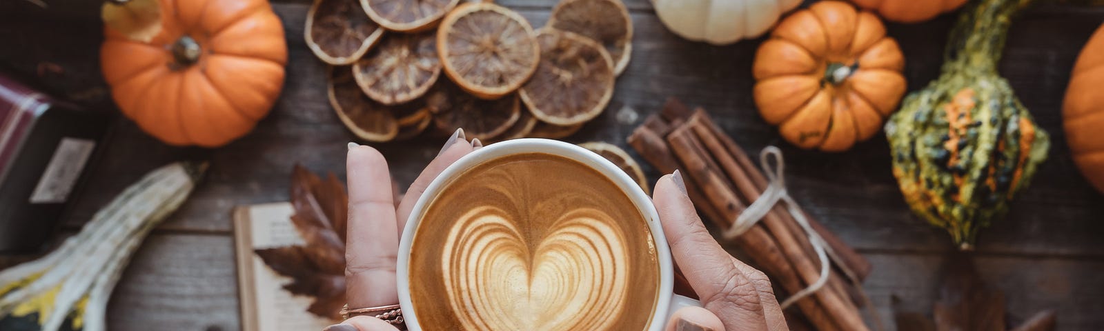 Hands half covered by the sleeves of a white jumper, cradle a mug of coffee topped with a heart pattern over a book sitting on a wooden table. An autumn theme is presented on the tabletop including pumpkins, chestnuts, preserved orange slices, and courgettes.