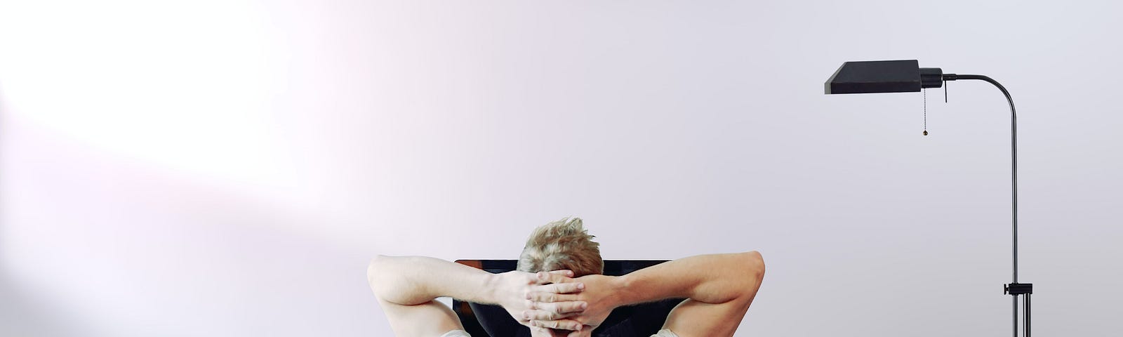 A blonde man sitting with his arms behind his head at a desk. There is a blank white wall with sunlight coming in on the lefthand side. On the right is a tall, skinny black lamp, and on the desk is an old-fashioned film camera, a small plant, and a monitor. The man’s posture makes him look irritated or tired.