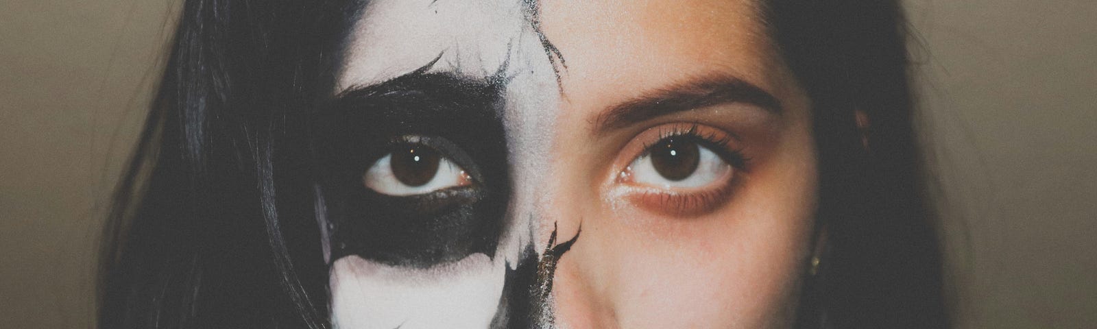 A closeup of the face of a young woman with dark hair, probably late teens or early twenties. Half her face is done up in skull / day of the dead makeup, the other half is regular makeup.