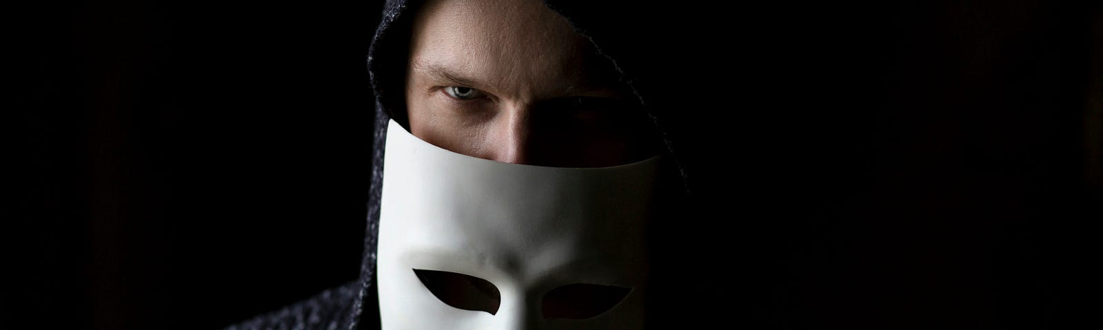 A man in a dark hoody peering over a white mask