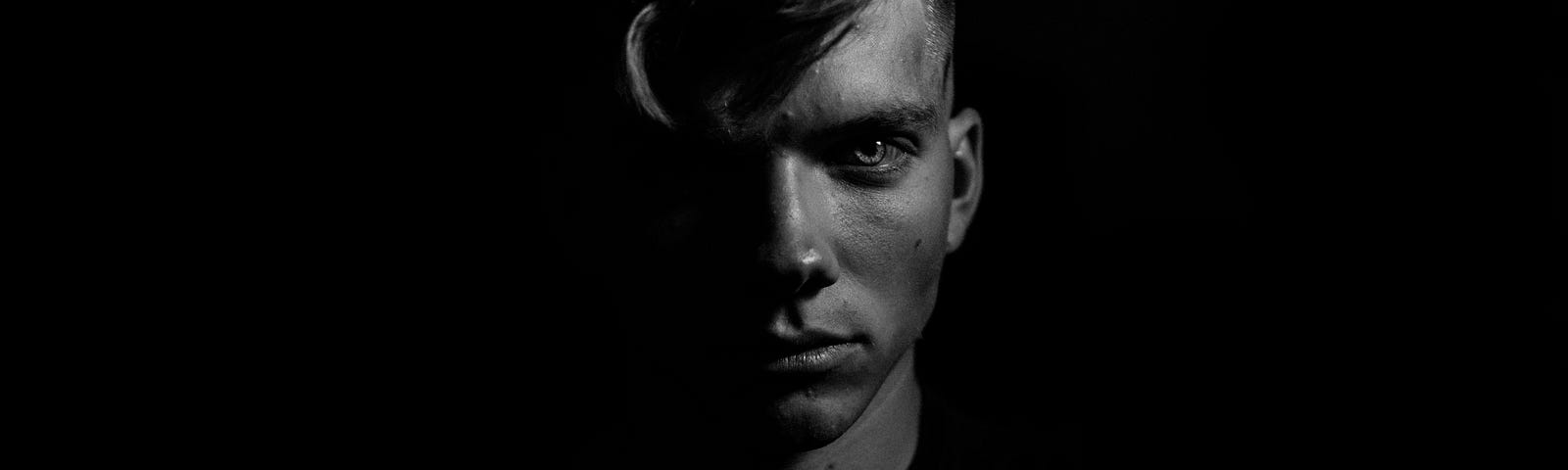 An image of an angular blond-haired male with an intense stare directed at the camera, half his face is cast in shadow, with a black backdrop.