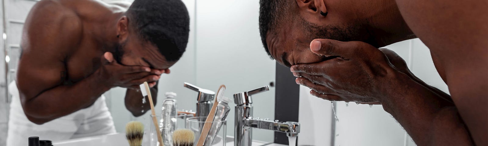 A muscular man is washing his face thoroughly in a white-tiled modern bathroom with a huge mirror.