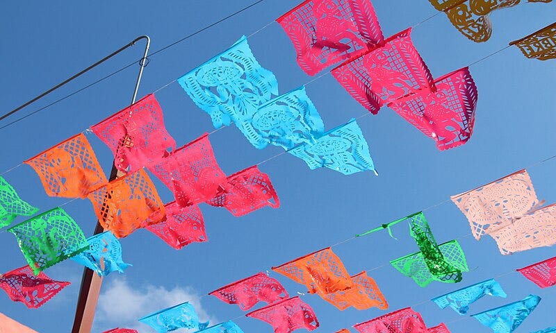 Against a bright blue sky, and colorful pieces of cut paper are hung along wires suspended in the air. Red, orange, blue, green and white, these decorative paper pieces flap in the wind like flags