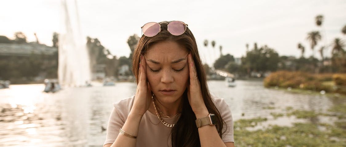 Woman sitting in front of body of water with her hands on her temples looking stressed