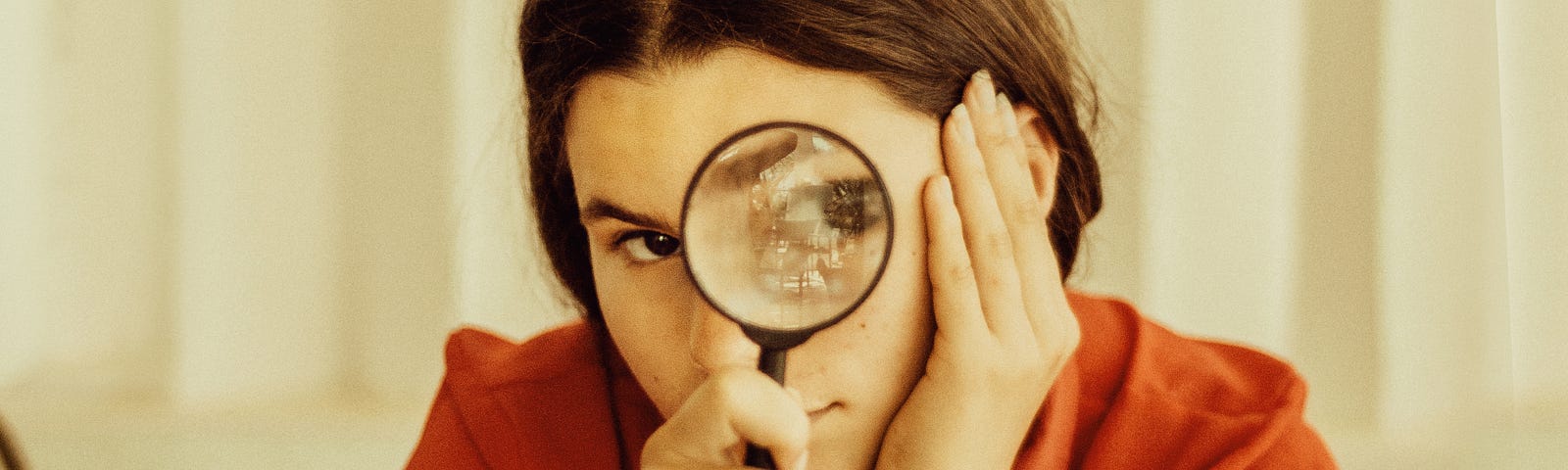 Woman sitting at a table with a magnifying glass held to her eye looking out at you with a book held open on the table with a half lemon on it