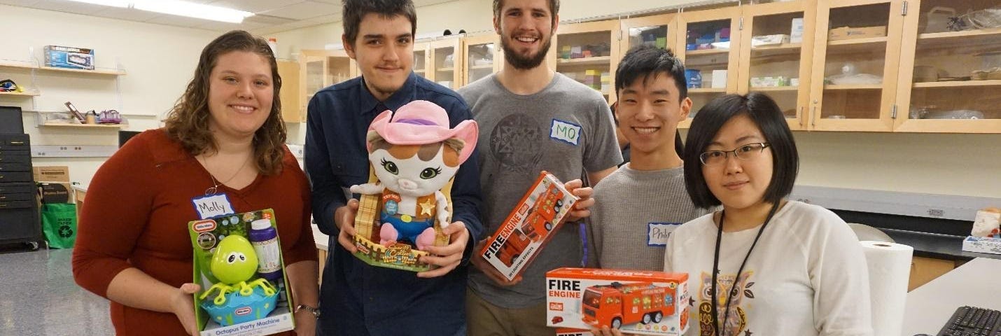Four people hold boxes with toys. In the background there are workbenches and drawers.