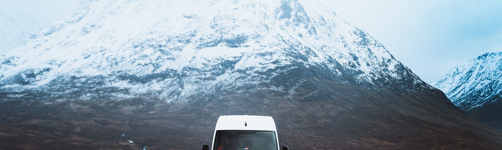 A high roof sprinter van driving down a road with snowy mountains behind it.