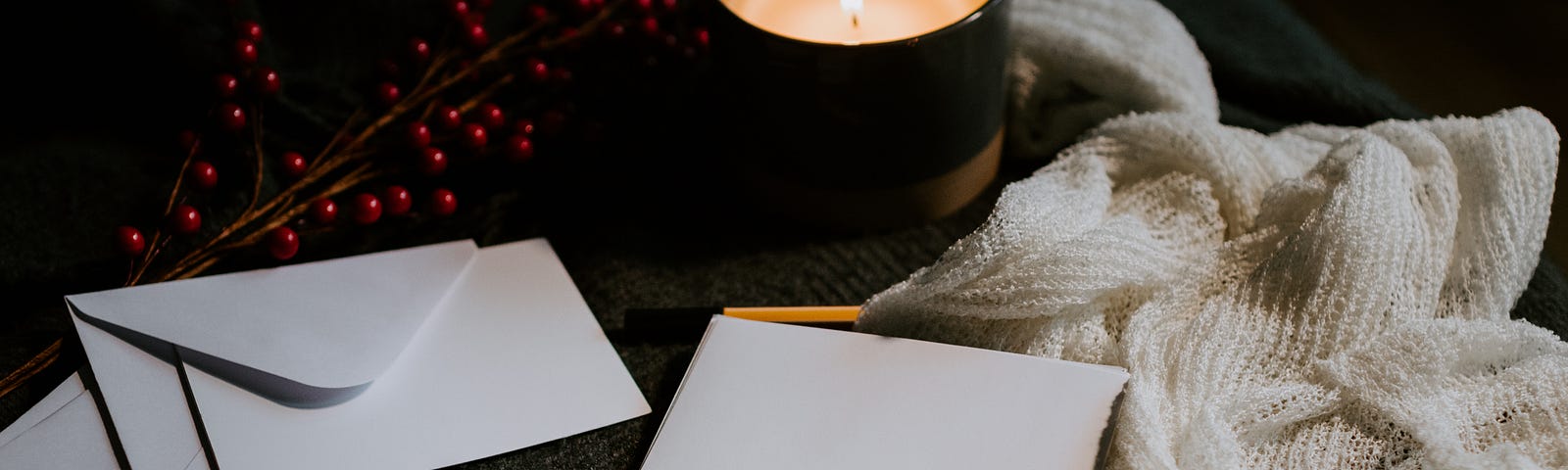 A candle surrounded by some stationary, a cloth, pen, and a branch with red berries