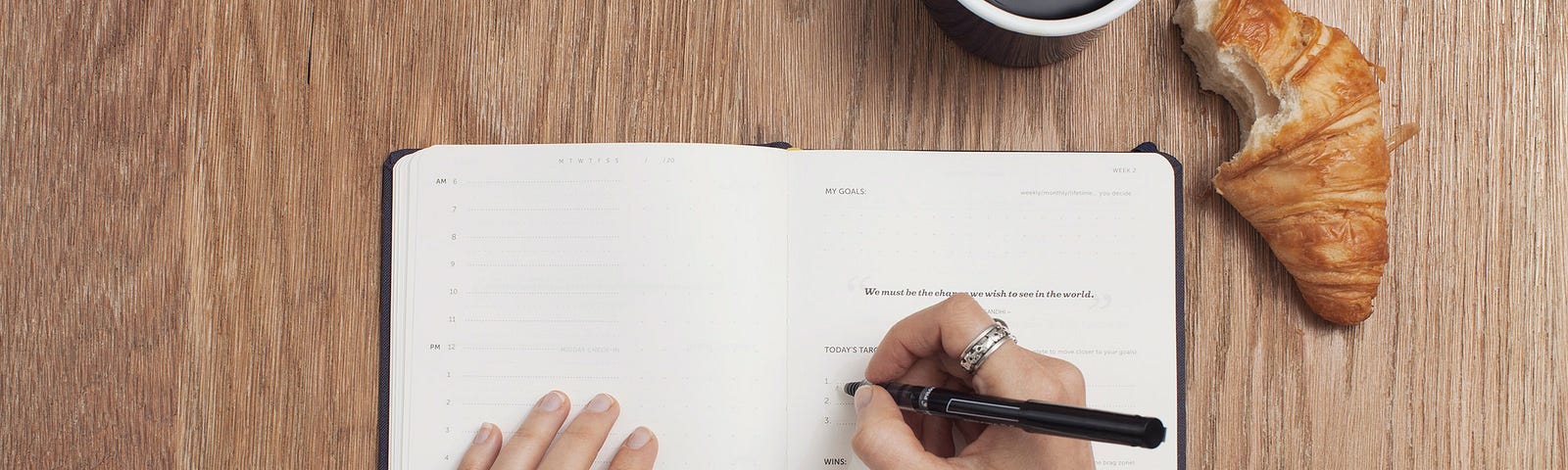 Photo of a person right-handed writing in a journal with white pages. Both hands are on the journal. To the top right of the journal, black coffee in a black mug with a croissant with one bite in it to the right of it.