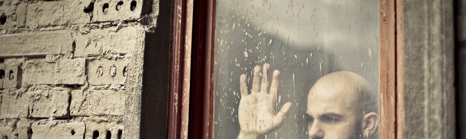 A bald man with a dark beard looks through a spattered window . His hand is pressed against the glass. Outside the casement, bricks of the living space seem unevenly laid.