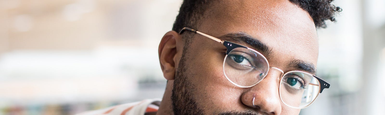 A black man in a red and white striped shirt sitting down, his hand under his chin. He’s wearing glasses and has a nose ring.