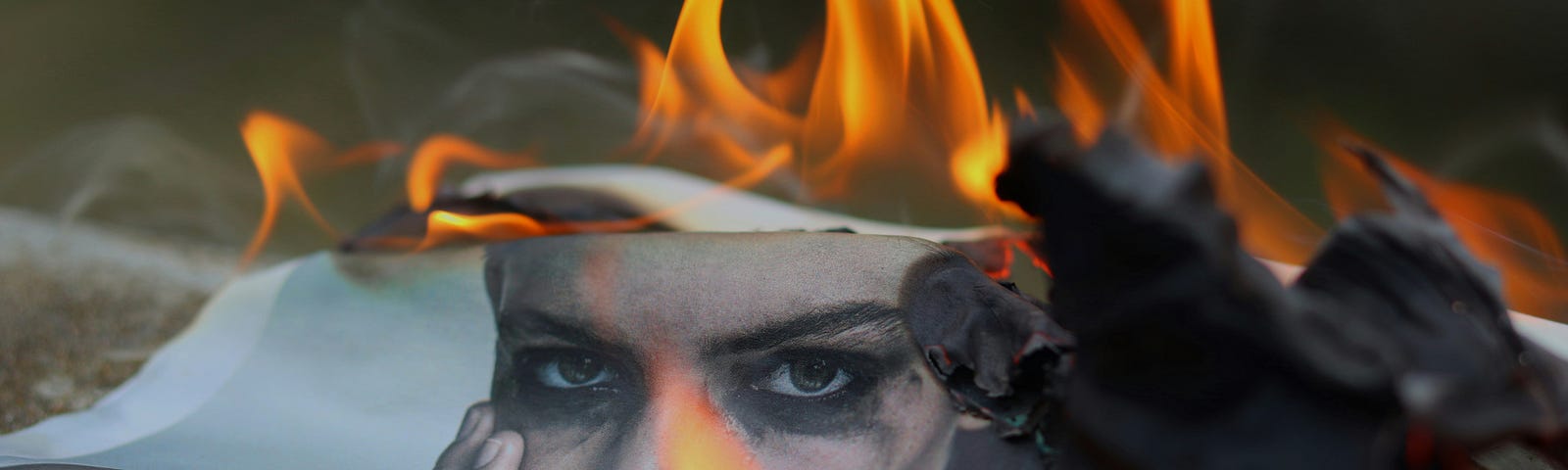 A photo of a woman’s face chars and curls as it burns on the ground. Her eyes stare into the camera.