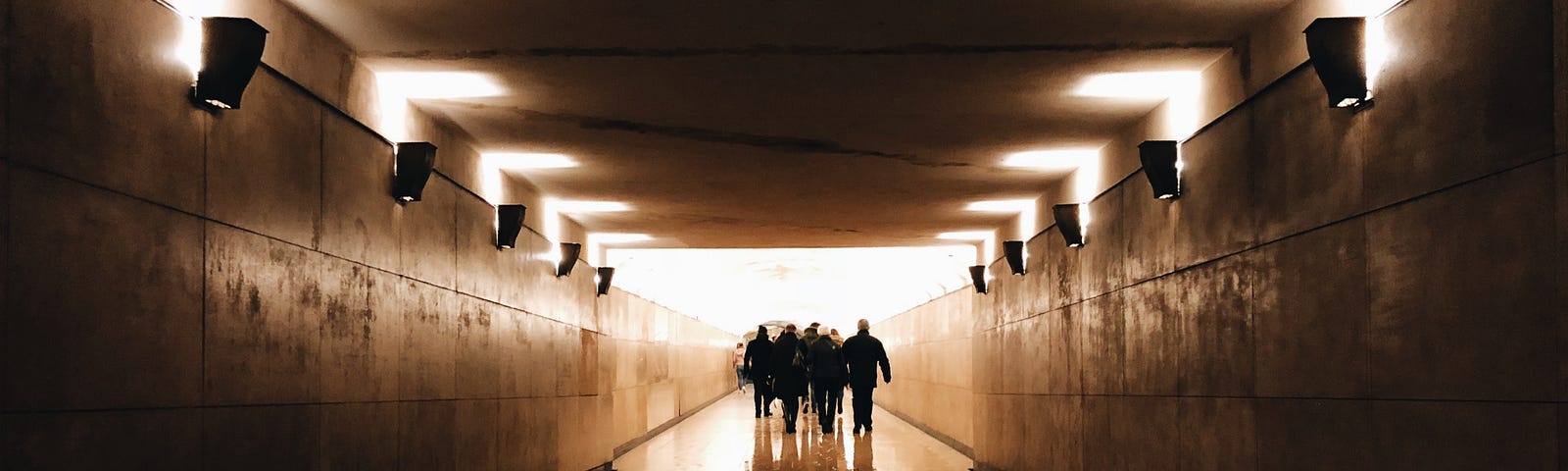 A group of individuals (in the distance) walks away from us in a walking tunnell with lights high on the side walls. While walking is often considered a leisurely activity, it can be an excellent form of exercise that offers numerous health benefits. A casual stroll can be enjoyable; adding intensity to your walk can turn it into a highly effective workout.