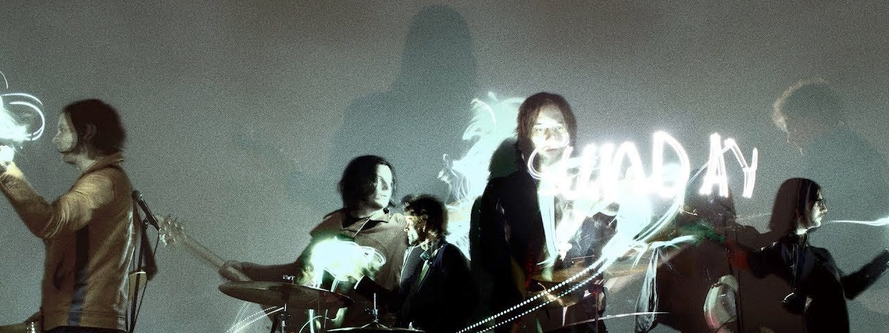 Shot from “Sunday Driver” music video; The Raconteurs band members playing their instruments and Jack White is shown in two places with the band and off to the left looking at white text that is written in the air around the band