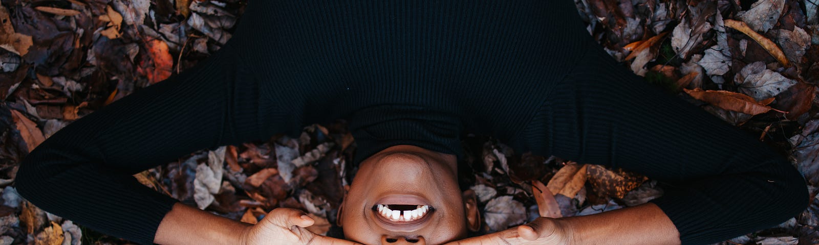 A black woman lays on a pile of leaves. She wears a black turtle neck and covers her eyes with both hands.
