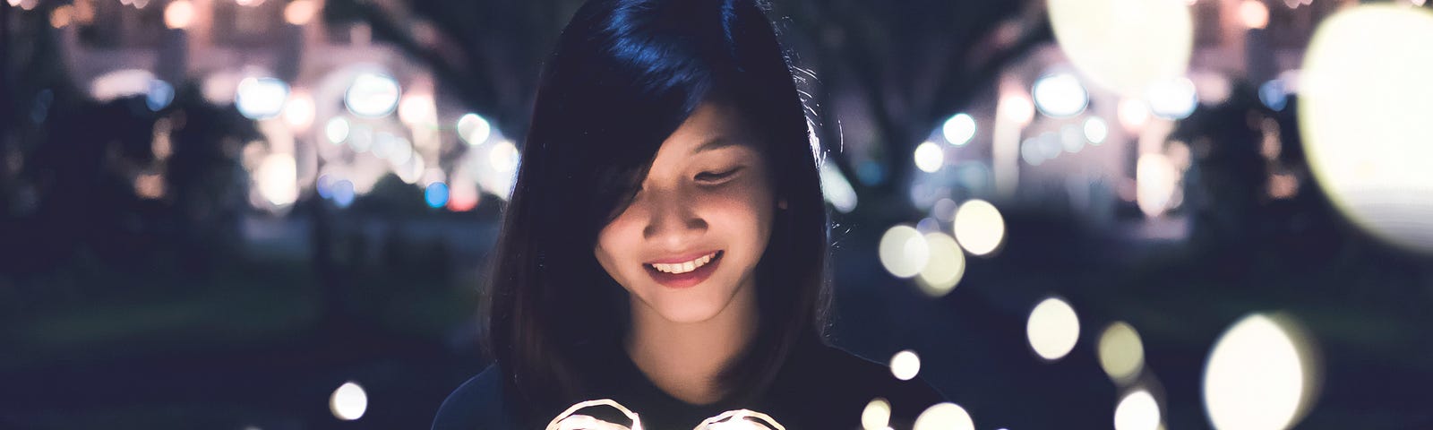 A young woman holding up a lighted white heart with lights around her in the background