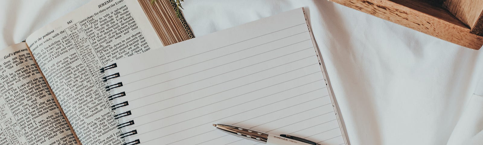 An instagram-worthy photo of a notebook. A beautiful pen rests on the surface of the lined page. Next to it, a book with tiny text rests open on the mattress of the bed, with a comfortable blanket draped around. Someone is taking notes for class.