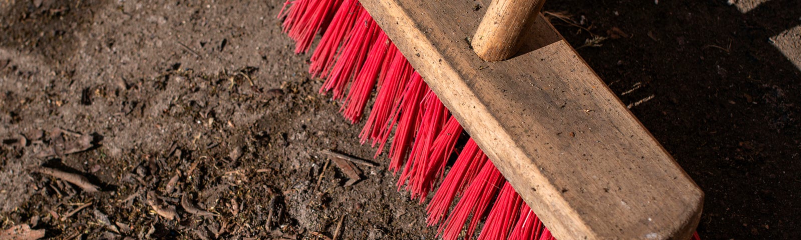 Close-up of a broom with red bristles sweeping dirt and debris on the floor, symbolizing the act of cleaning up and removing unnecessary items.