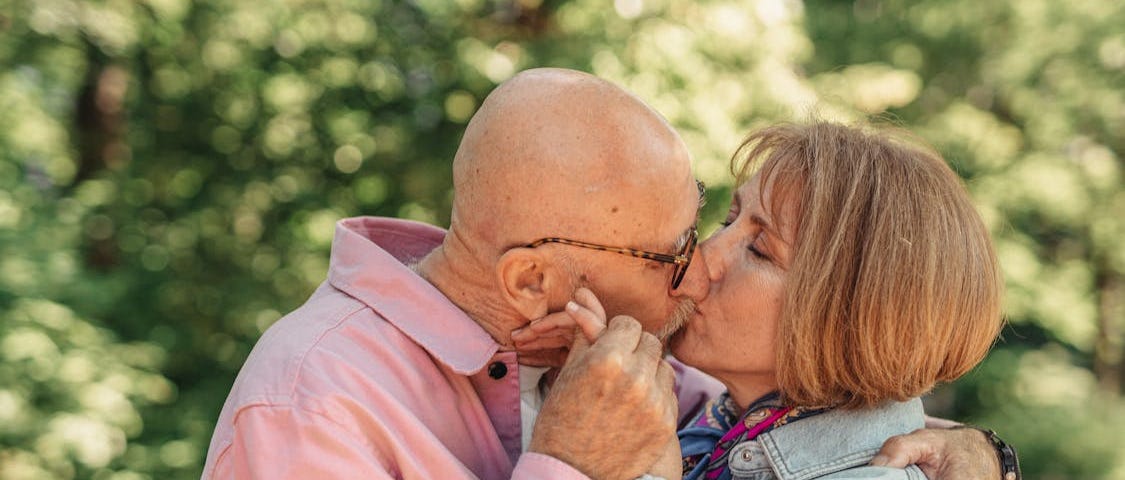 A bald light-skinned older man with glasses is on the left, wearing a pink button-down shirt, kissing, embracing, and holding the hand of a light-skinned older woman with chestnut hair styled in a bob, wearing a light denim button-down shirt and a colorful scarf. They’re outside amongst trees in the sunlight.